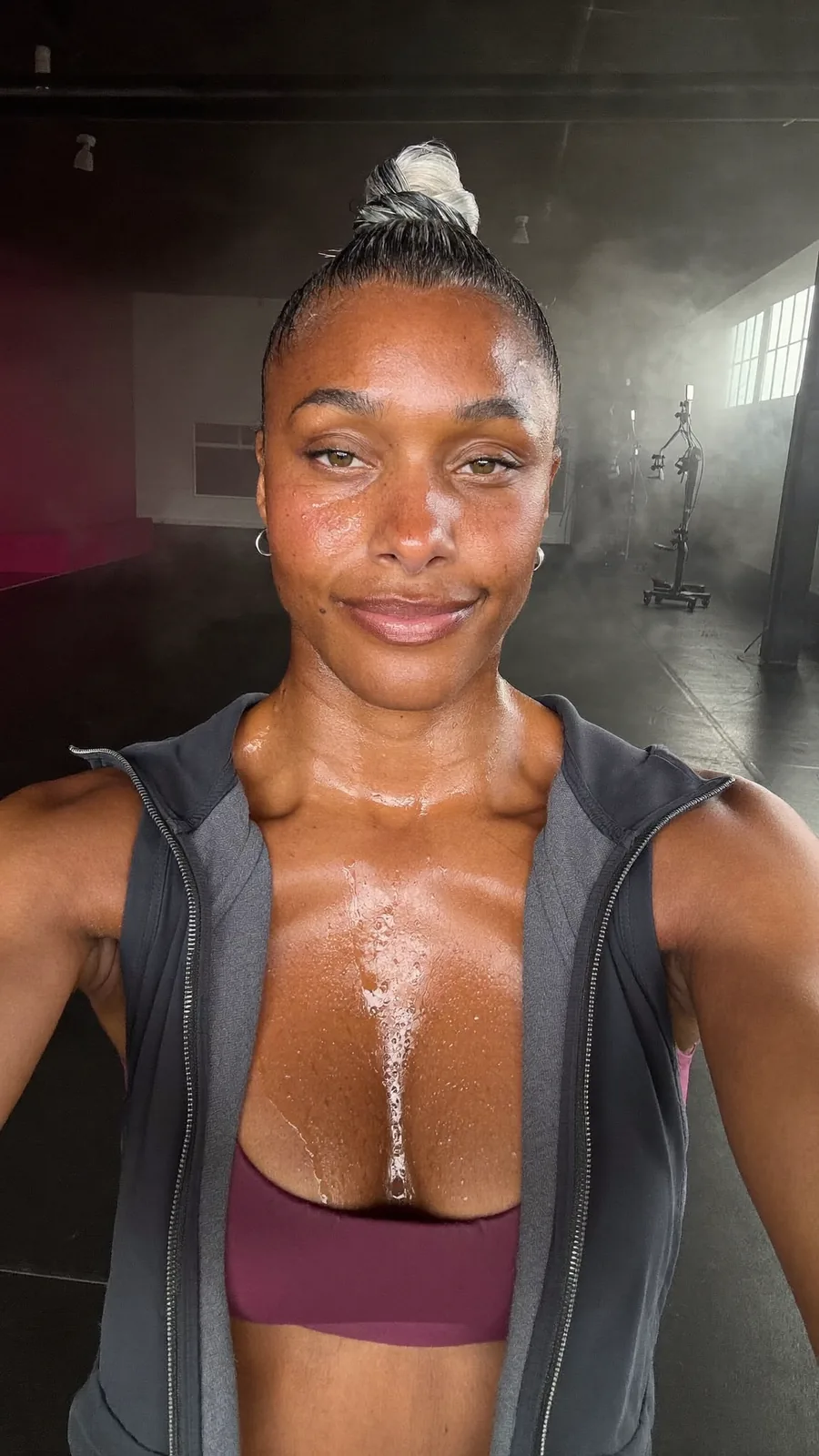 The head coach, a woman with a high bun streaked with grey, in an open charcoal jacket, with a calm, dry smile in the dim studio