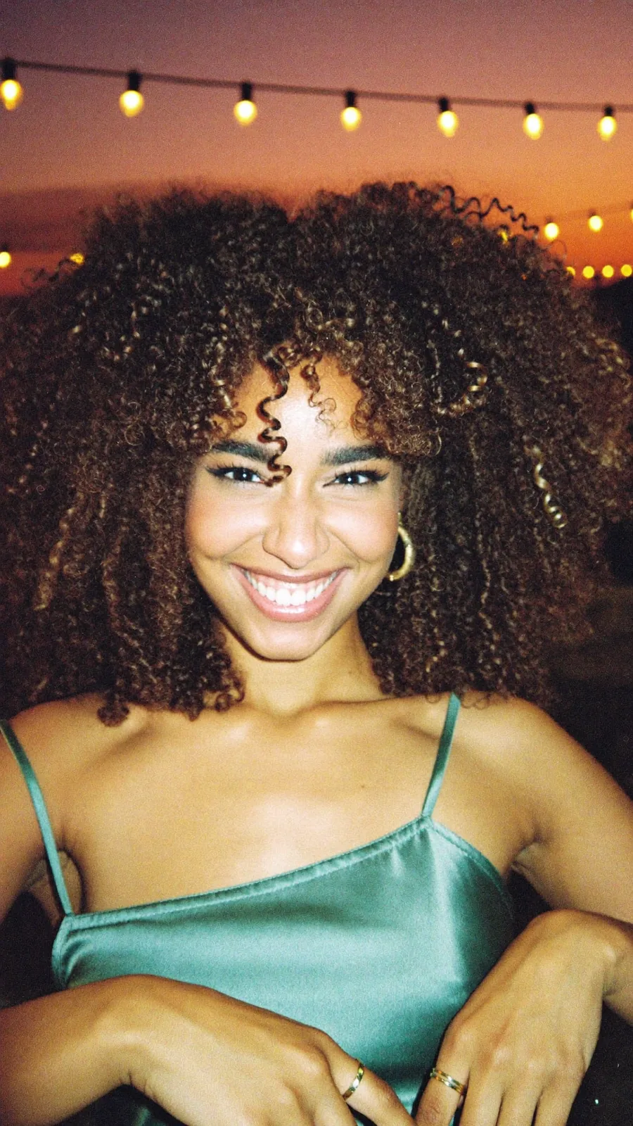 The same young woman in a teal satin top, grinning into a direct-flash photo at a rooftop party
