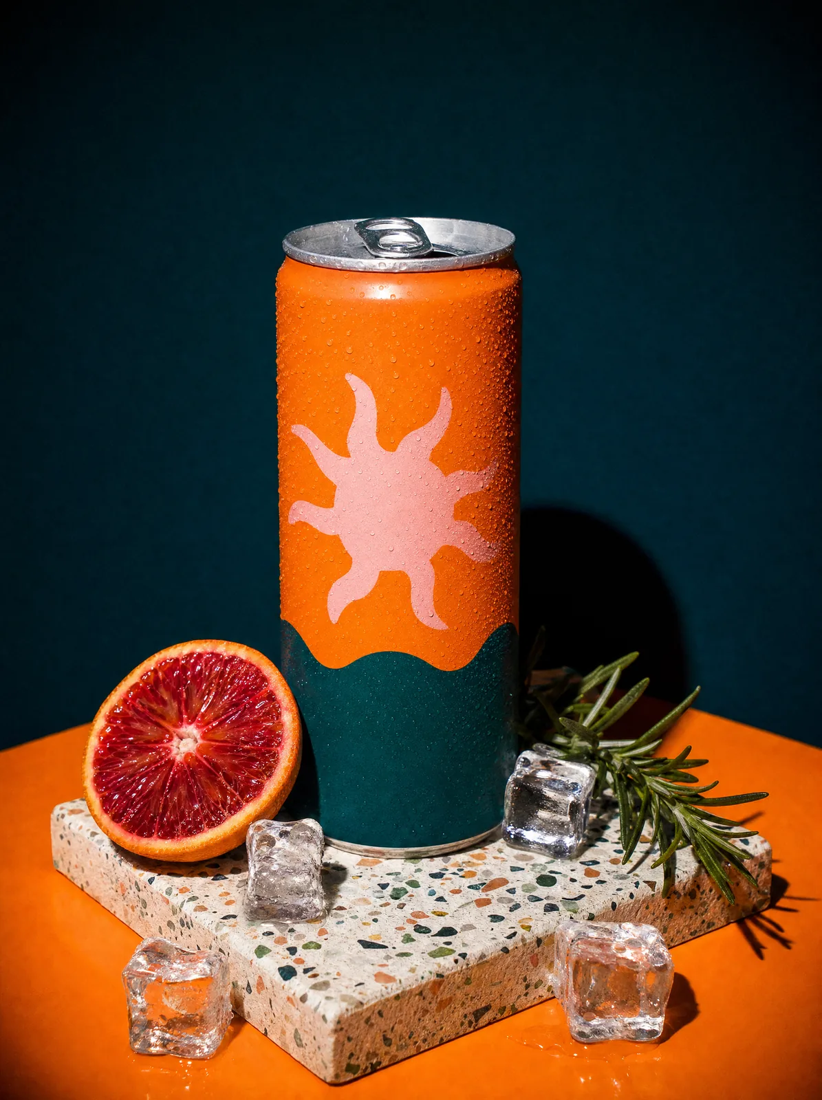 A slim tangerine can with a pale-pink sun graphic and wavy teal band on a terrazzo block with blood orange, rosemary and ice